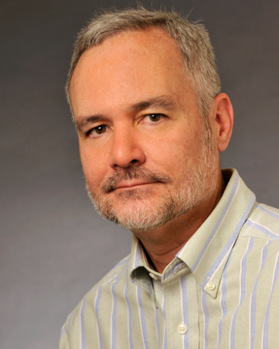 A man wearing a light-colored collared shirt with blue stripes looks at the camera.